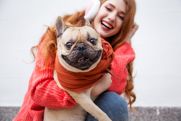 Beautiful teen redhear girl holding the dog against street wall. Young girl wearing sweater, jeans. dog wearing scarf.