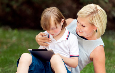 Young cheerful mother hugging her son using a tablet on a park lawn