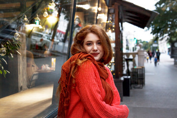 Fototapeta premium Young beautiful woman walking at city street at cool christmas day in garland bacground. Wearing warm oversize sweater and scarf.