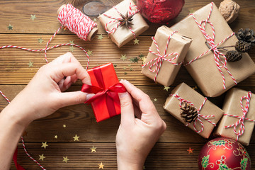 Female hands holding, wrapping Christmas gift box. Group of gift boxes wraped in craft paper, red baubles, glitter over wooden tables. Chritsmas flat lay background.