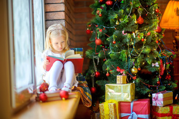Fototapeta na wymiar Cute girl in a white dress sitting on a saucer is reading a book, next to a festive tree and gifts. Happy children. Cristmas presents.