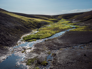 Small stream from melting ice with Lush vivid green wet moss in black lava hills in Landmannalaugar mountain. Fjallabak Nature Reserve in Highlands of Iceland, summer blue sky.