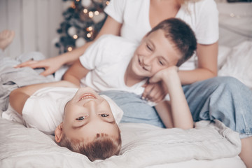 Fototapeta na wymiar Happy loving family. Beautiful young mother and her two kids child boys playing, laughing and hugging in white Scandinavian style bedroom against the background of the Christmas tree