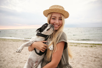 Portrait of happy young blonde female with long hair looking at camera with charming smile while keeping her beloved dog in hands, posing over seaside on gray cloudy day