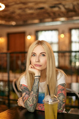 Portrait of beautiful blonde long haired female with tattooes leaning chin on raised hand and looking a camera with calm face, posing over restaurant interior in casual clothes