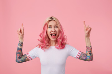 Overjoyed pretty young pink haired woman with tattoos pointing upwards with forefingers and opening...