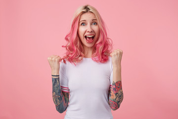 Portrait of young attractive tattooed woman with pink curly hair looking at camera joyfully and raising happily fists, standing over pink background in casual clothes