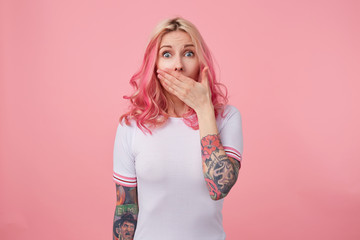 Indoor photo of open-eyed tattooed young female posing over pink background in casual clothes,...