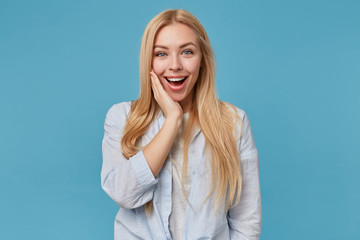 Joyful attractive young long haired blonde lady leaning her chin on raised palm, standing over blue background in casual clothes, smiling happily and looking sincerely to camera