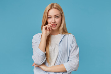 Fototapeta premium Horizontal photo of positive blonde long haired woman in casual clothes posing over blue background, keeping index finger on underlip and smiling to camera sincerely