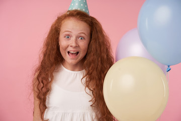 Fototapeta na wymiar Portrait of joyous redhead girl with long curly hair in white dress and birthday cap being excited and surprised to get birthday present, happily looking in camera over pink background