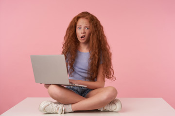 Photo of curly female teenager with long red hair grimacing over pink background, sitting with crossed legs in casual clothes, holding laptop while looking at camera with wide eyes and mouth opened