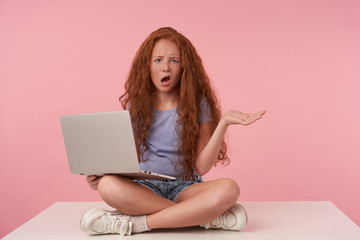 Studio shot of redhead curly girl with long hair looking at camera with confused face and holding laptop on legs, raising palm with pout, posing over pink background in blue t-shirt and jeans shorts
