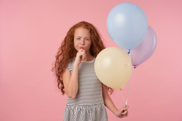 Charming redhead curly long haired girl in casual clothes standing over pink background, looking thoughtfully to camera and holding chin with raised hand, keeping air balloons in her hand