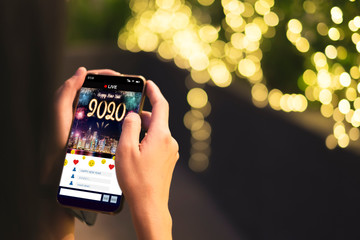 Woman Hand holding mobile see live streaming countdown to Happy new year 2020 with fireworks over cityscape on screen with light bokeh background,Digital social media lifestyle