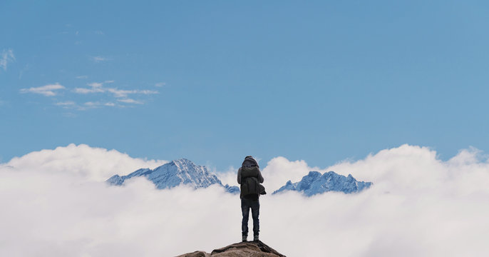 a man with backpack standing on mountain top, with snow mountains over clouds. Success, achievement and life goal concept