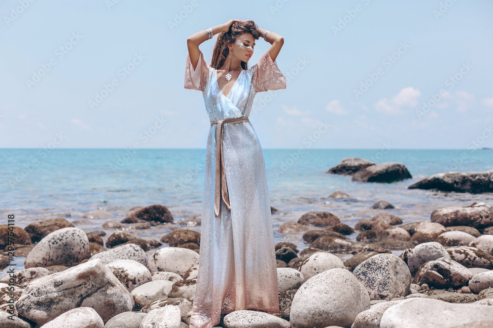 Wall mural beautiful young fashionable woman in elegant dress posing at the stone beach at sunset