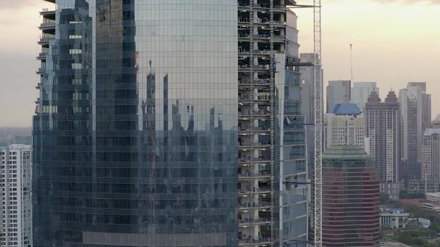 pull-back zoom out aerial view of construction, almost finished tall building cover with glass. Crane, City Reflection of Sunset
