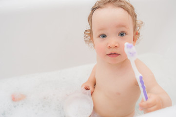 Funny happy baby with curly hair taking bath and brushing her teeth. A cute baby girl is happy in the bathroom. Cute Toddler giving satisfied smile from the bathtub. Small child bathes in a bathroom