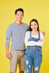 Positive young Vietnamese man is striped shirt hugging his pretty girlfriend wearing denim overall