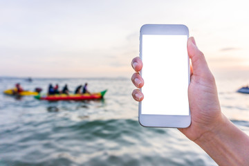 Mockup image of a hand holding and showing white mobile phone with blank desktop screen in front of blur the sea sky and person play banana boat background