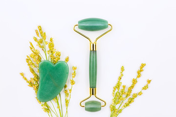 Gua sha stone and jade roller still life arranged with golden wildflowers on white surface /...