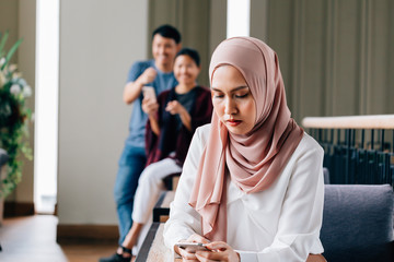 Islamic Southeast Asian lady in hijab browsing smartphone while being abused by blurred people of...