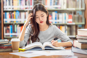 Asian young Student in casual suit reading and doing homework in library of university or colleage with various book and stationary on the wooden table over the book shelf background, Back to school