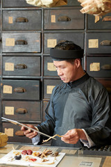 Chinese medicine practitioner following list on tablet computer when gathering dry ingredients for recipe