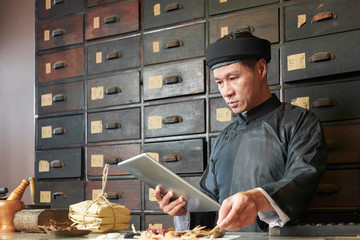 Serious traditional medicine practitioner reading old book on tablet computer when gathering...