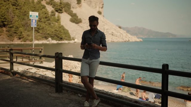 Relaxed Man Looking And Texting On Mobile Phone In Vacation Near Sea.Male Chatting With Friends.Man Using Mobile Phone On Vacation In Sunny Day.Man Having Chat On Smartphone At Mediterranean Beach.