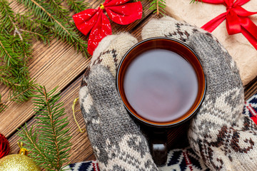 hands in mittens holding cup of tea close up on the brown brushed wooden fur pine branches gift toys background. Autumn leaves  hygge, fika, lagom, cozy, warm, relax holidays concept