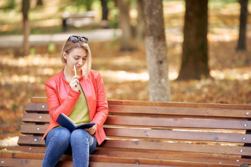 Obraz premium young woman is reading a book while sitting on a bench in a city park. Autumn weather, golden leaves.
