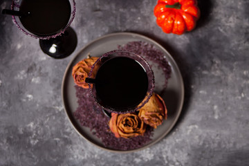 Two glasses with black cocktail, dried roses, pumpkin for Halloween party on the dark background
