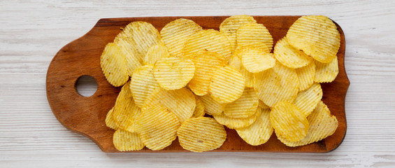 Salted wavy potato chips on a rustic wooden board on a white wooden table, top view. Overhead, flat lay, from above. Close-up.