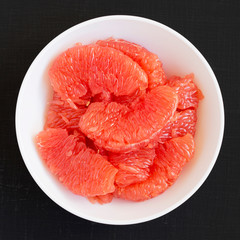 Red grapefruit slices in a white bowl on a black surface, overhead view. Flat lay, top view, from above. Close-up.