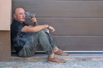 Fototapeta premium A middle-aged man is resting on the sidewalk and smoking pipe. Portrait of caucasian bald man, outdoors, close up. Bad habits, addiction. Unhealthy lifestyle concept.