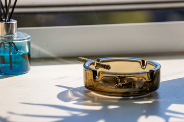 A marijuana blunt joint is burning in a glass ashtray near a window at home.
