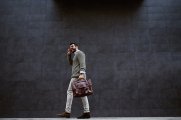 Full length of stylish caucasian designer in gray sweater holding leather bag while passing by gray wall.