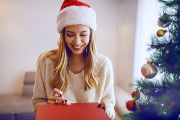 Beautiful caucasian blonde woman with santa hat on head smiling and holding christmas gift while standing in living room. In foreground is decorated fir tree.