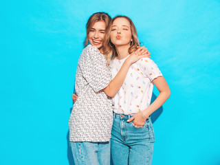 Two young beautiful smiling blond hipster girls in trendy summer colorful T-shirt clothes. Sexy carefree women posing near blue wall. Positive models having fun