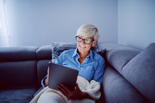 Beautiful Smiling Blonde Caucasian Senior Woman Sitting On Sofa In Living Room And Using Tablet For Internet Surfing.