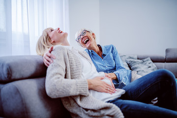 Happy senior woman sitting next to her pregnant daughter on sofa in living room and touching her...