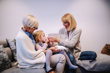 Sick granddaughter sitting in granny's lap and blowing nose while her mother helping her. Living...