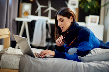 Attractive caucasian brunette in blue and black sweater lying on stomach in bed and typing on...