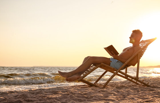 Young Man Reading Book In Deck Chair On Beach