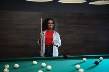 Fototapeta premium Young African-American woman with cue near billiard table indoors