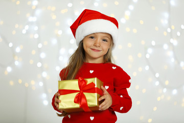 Happy little child in Santa hat with gift box against blurred festive lights. Christmas celebration