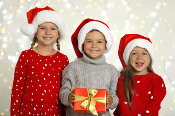 Happy little children in Santa hats with gift box against blurred festive lights. Christmas celebration