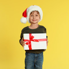 Cute little boy in Santa hat with Christmas gift on yellow background